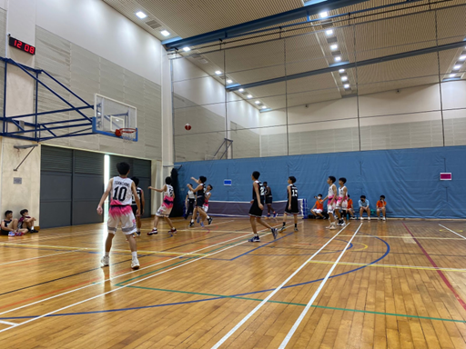 Photograph of an indoor basketball game with players from two teams wearing black and white-pink uniforms actively engaged near the hoop. The court features wooden flooring, a visible scoreboard showing 12:08, and spectators seated along the wall.